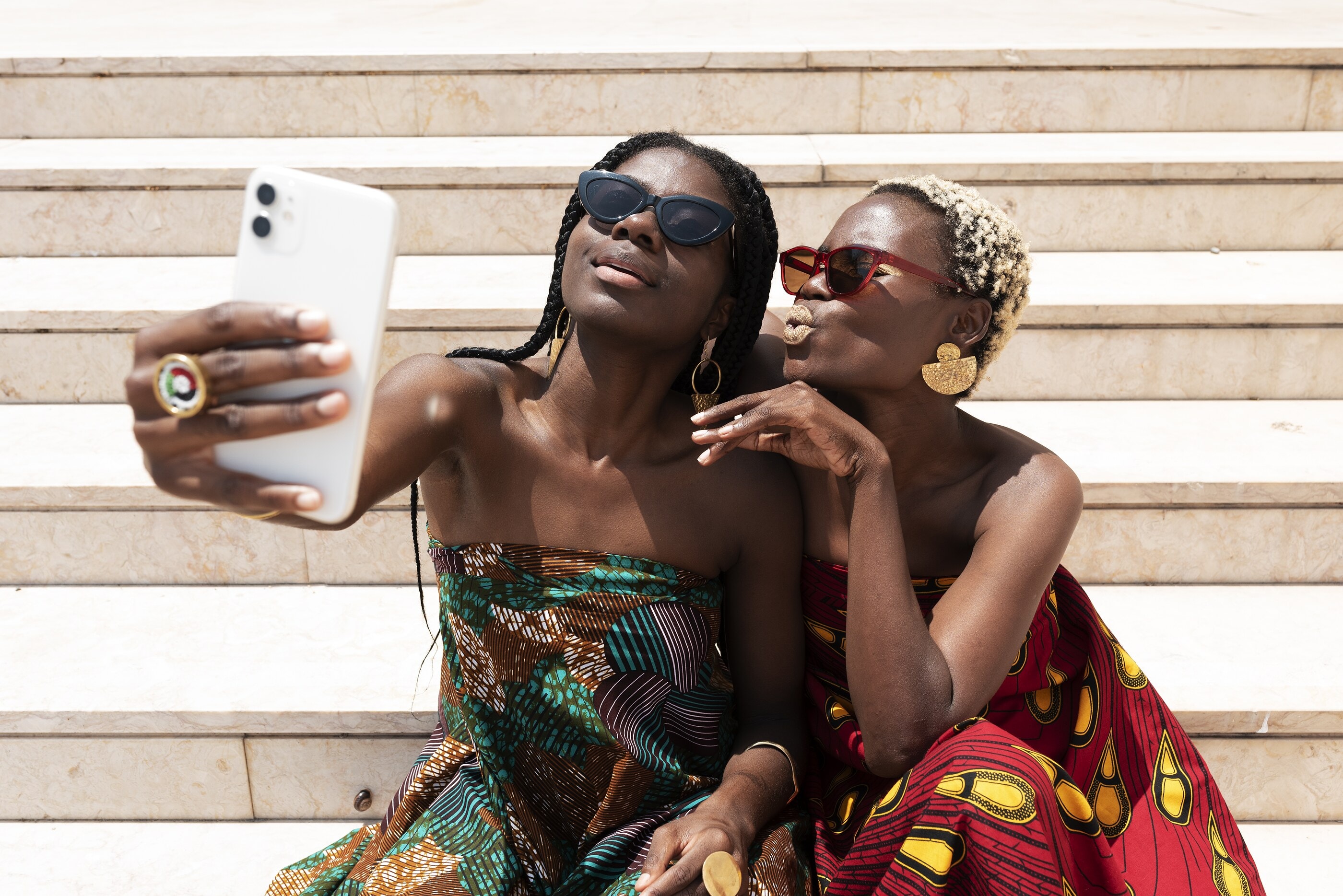 Two friends in wax-print outfits taking a selfie on sunlit steps