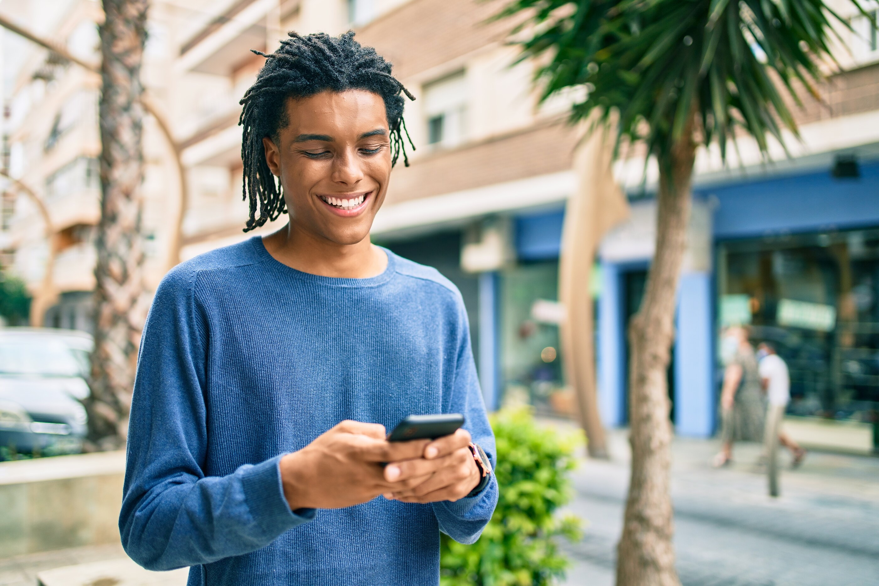 A young man smiling at a message on a sunny street lined with palms